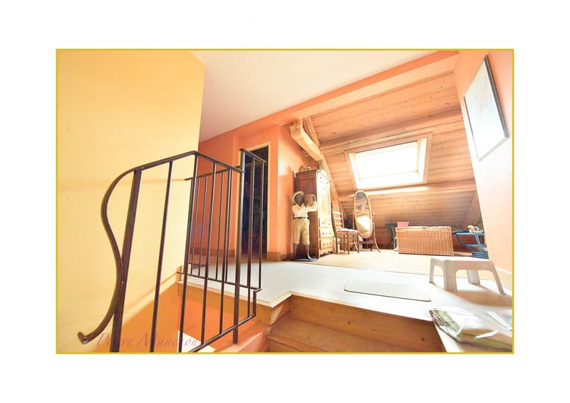 A cozy, well-lit room with wooden walls and a slanted ceiling. Features include a metal railing, a small table, a wicker basket, a mirror, a statue, and a cabinet.