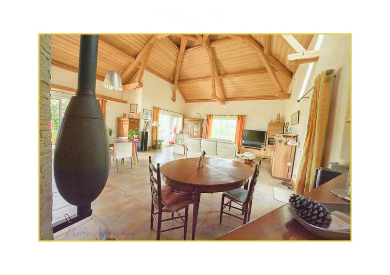 A spacious living and dining area with exposed wooden beams, a large stone fireplace, a round wooden dining table with chairs, a white sofa, a television, and wooden cabinets.