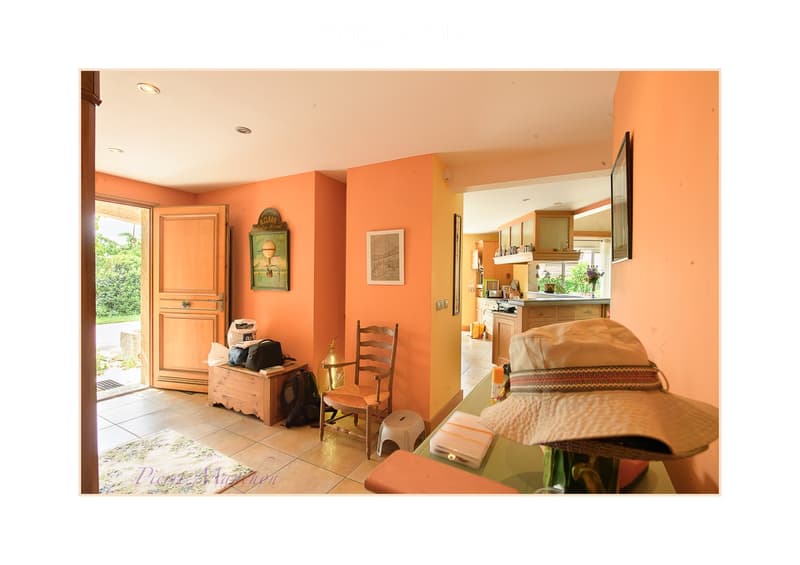 Brightly colored hallway with peach walls, a wooden chair, a small table with bags, a hat on a stand, and an open doorway leading to a kitchen area with cabinets and appliances.