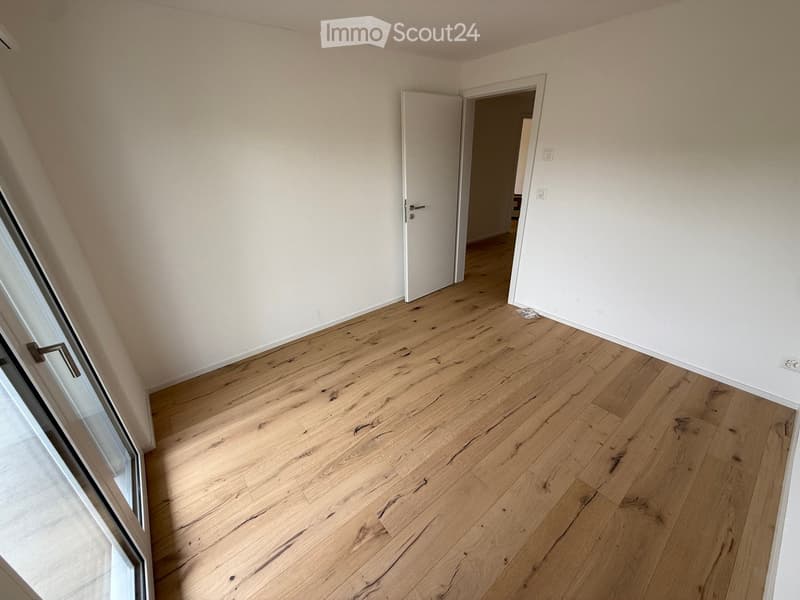 Empty room with light wooden flooring and white walls, featuring a door leading to another room and a large window with a view.