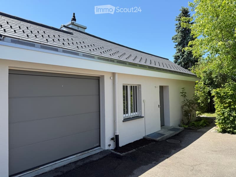 Single-story house with a grey garage door, white exterior walls, and a small window with bars. The roof is grey with a patterned design, and the house is surrounded by greenery.