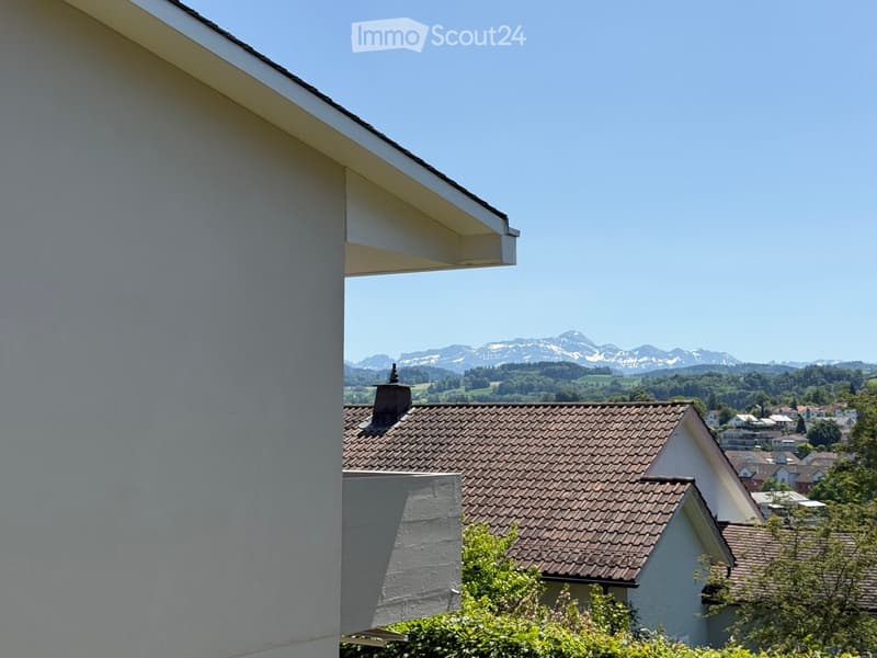 View from a balcony showing a residential area with tiled roofs, trees, and distant mountains under a clear blue sky.