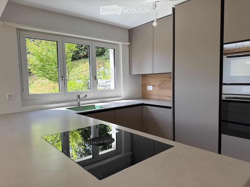 Modern kitchen with white countertops, grey cabinets, a sink with a chrome faucet, and a built-in oven. Large windows provide natural light and a view of greenery outside.
