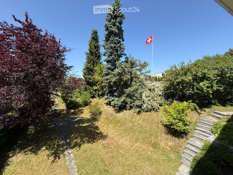 A well-maintained garden with a stone pathway, grassy area, and various trees and shrubs. A Swiss flag is visible on a pole in the background.