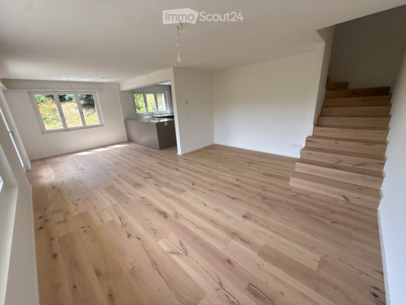 Empty room with light wooden flooring, white walls, and a staircase leading upwards. A small kitchen area with cabinets and a sink is visible in the background, along with windows letting in natural light.