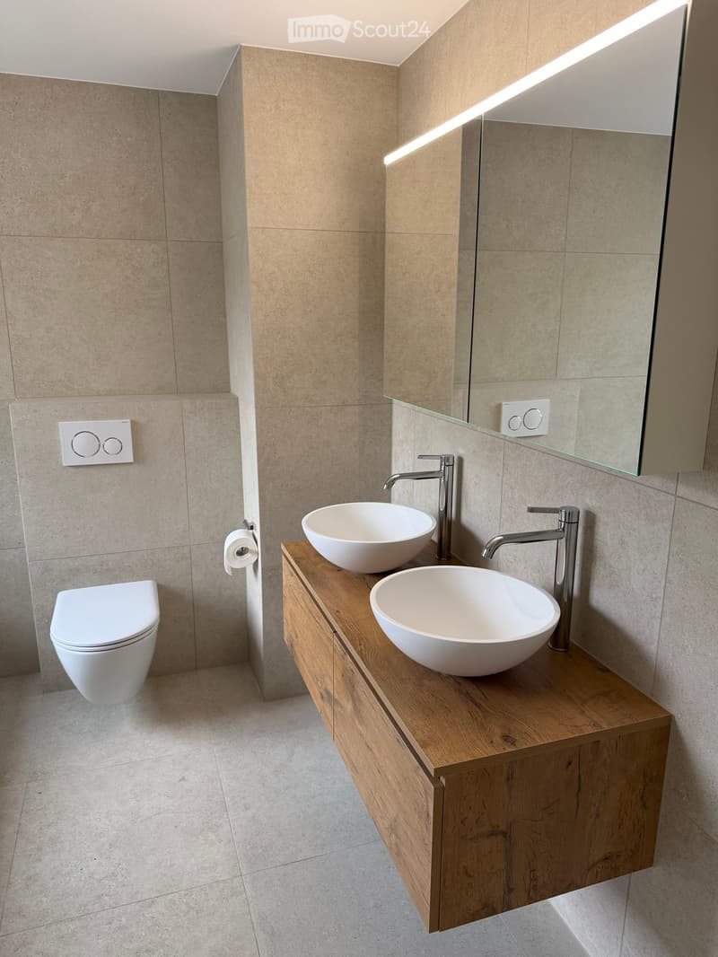 Modern bathroom with a wooden vanity, two white sinks, sleek chrome faucets, a large mirror, and a toilet.