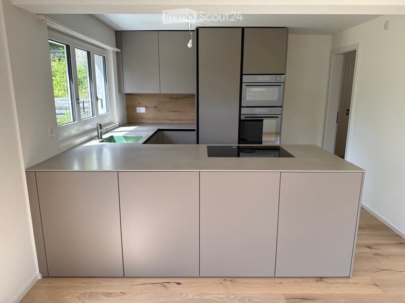 Modern kitchen with grey cabinets, a central island, a sink, and an oven. The space features large windows and a wooden backsplash.