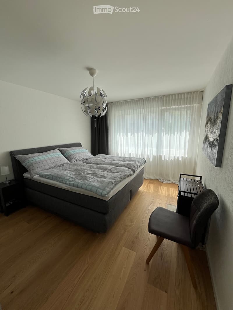 Modern bedroom with wooden flooring, a large bed with grey bedding, a nightstand with a lamp, a chair, and a decorative wall light fixture.