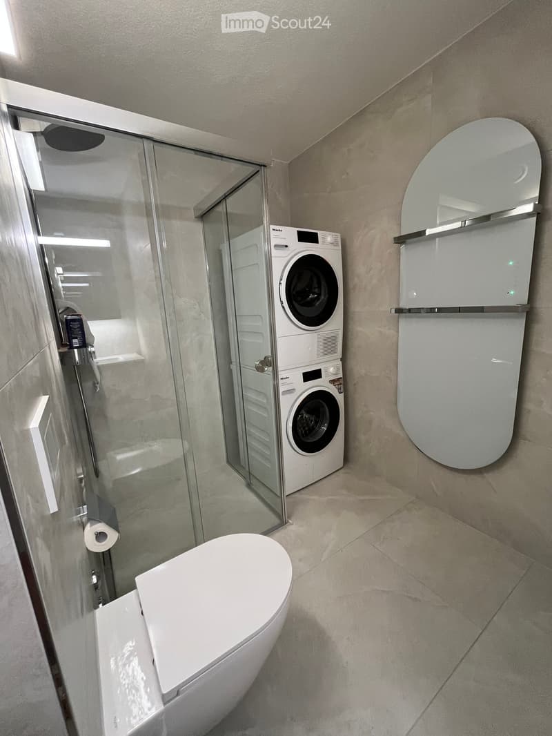 Modern bathroom with a toilet, washing machine, glass-enclosed shower, and a large round mirror above the toilet.