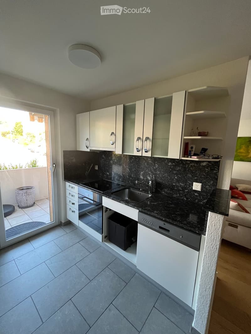 Modern kitchen with white cabinets, granite countertop, stainless steel sink, and built-in appliances including an oven and dishwasher. The space features tiled flooring and an open doorway leading to a balcony area.