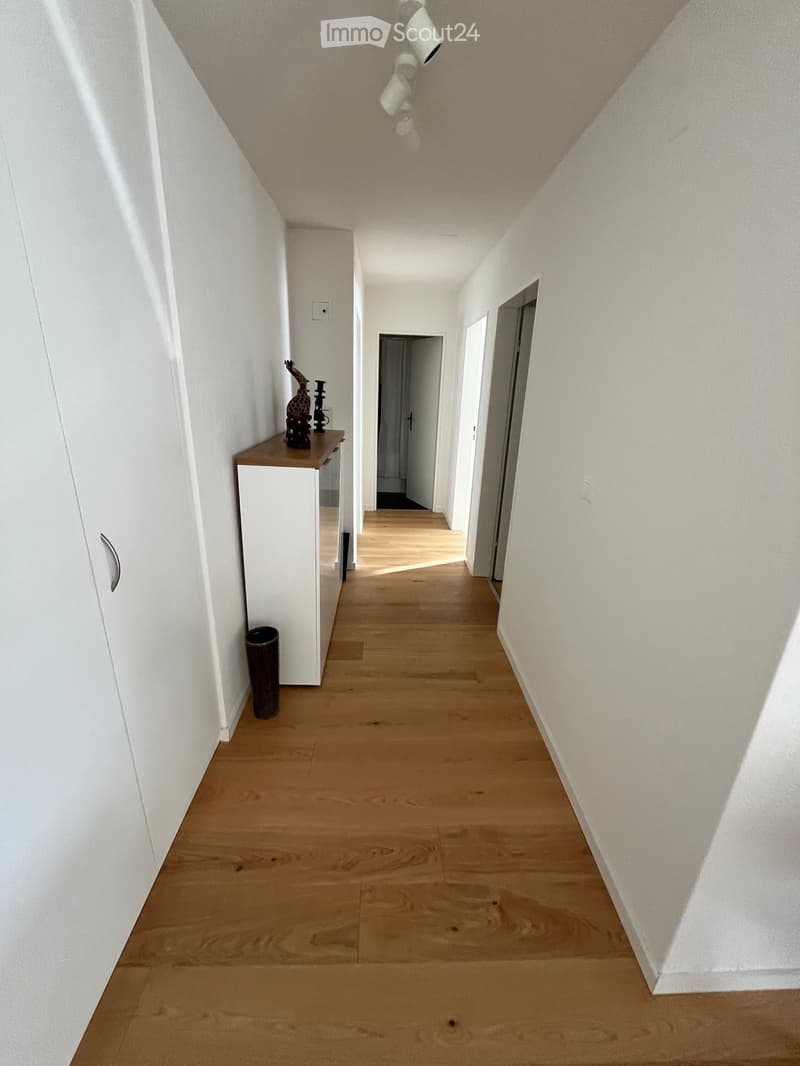 A long, narrow hallway with light wooden flooring and white walls, featuring a small wooden console table with decorative items and a trash can on the left side.