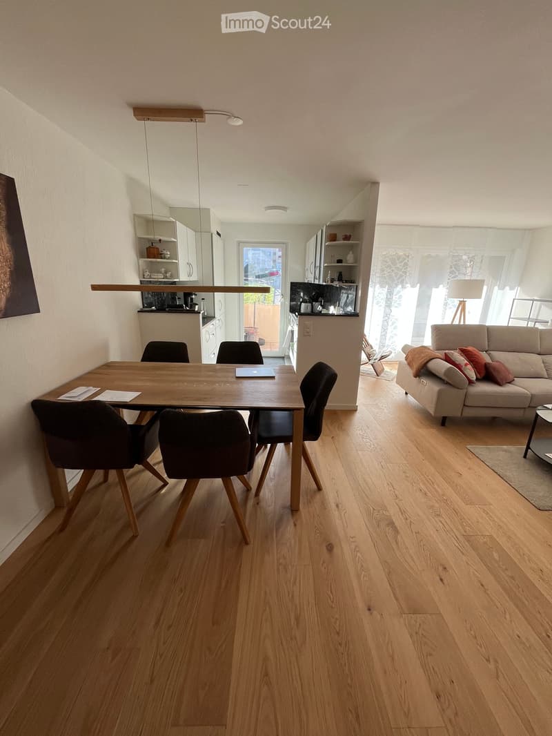 Open-plan living and dining area with light wooden flooring, a sofa with cushions, a dining table with chairs, a laptop on the table, and a modern kitchen area with cabinets and appliances.
