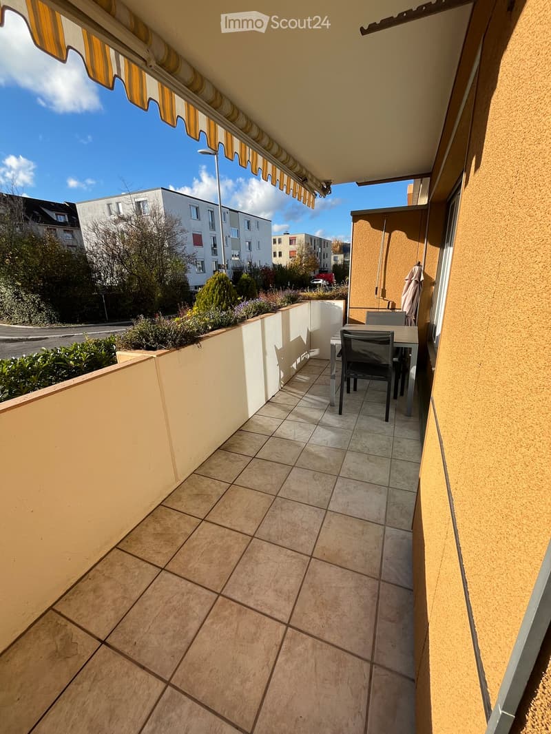 Balcony with tiled floor, small table and chairs, covered by a striped awning, with a view of neighboring buildings and greenery.