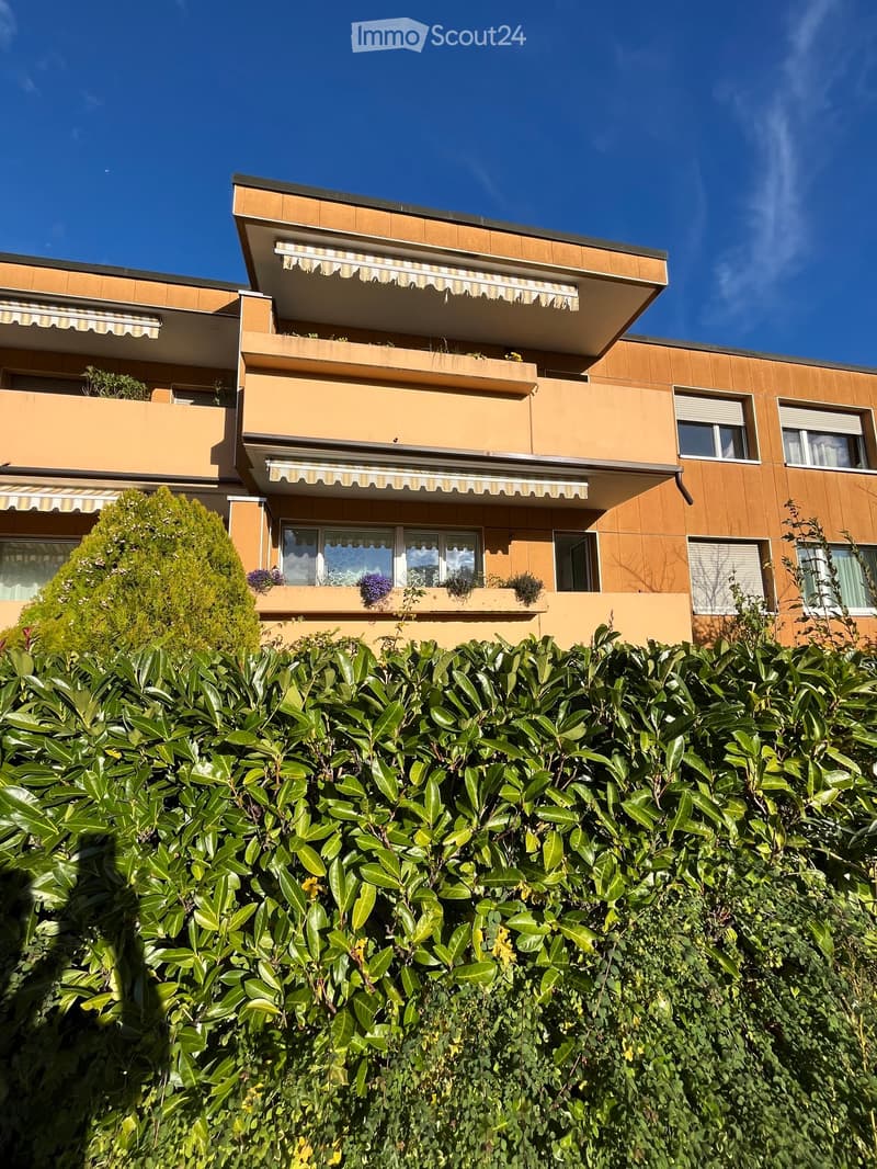Modern multi-story building with balconies, large windows, and striped awnings, surrounded by lush greenery.