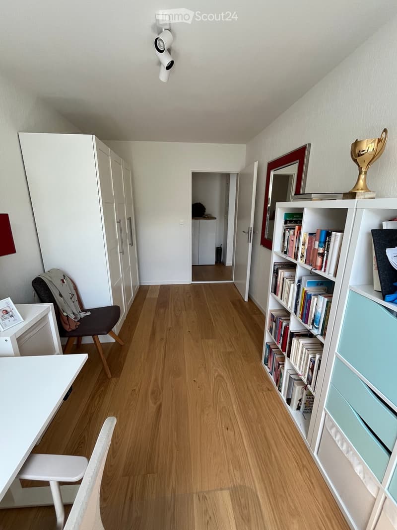 A well-lit room with wooden flooring, a white table, a white chair, a dark-colored chair with a jacket draped over it, a bookshelf filled with books, a mirror, and a small cabinet.