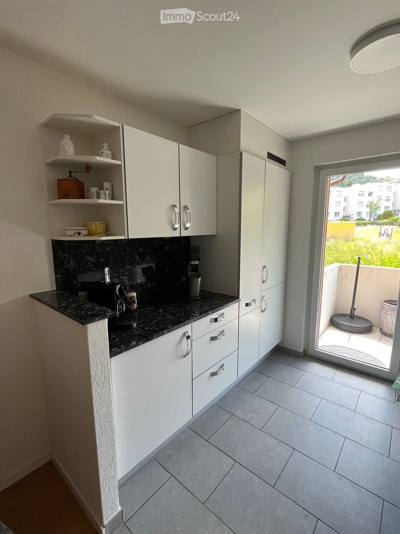 Modern kitchen with white cabinets, granite countertop, sink, coffee maker, and open shelving with decorative items.