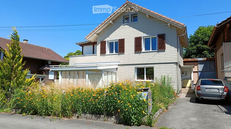 Two-story house with white siding, red-brown shutters, and a small balcony. The house has a well-maintained garden with yellow flowers and a parked car in the driveway.