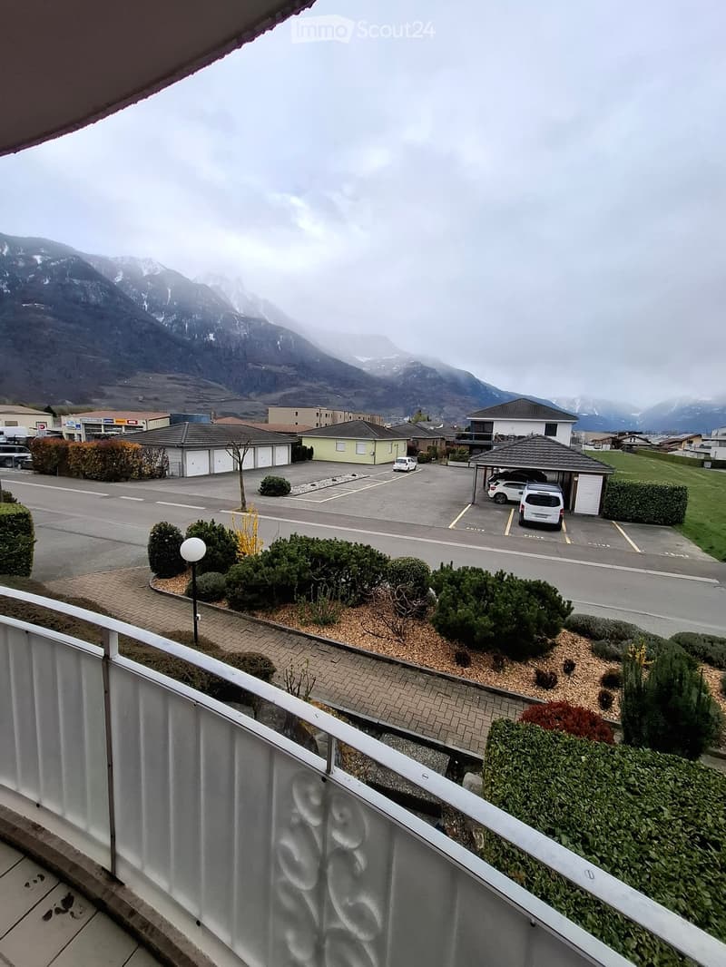 Balcony overlooking a parking lot with several cars, surrounded by neatly trimmed bushes and shrubs, with distant mountains visible in the background.