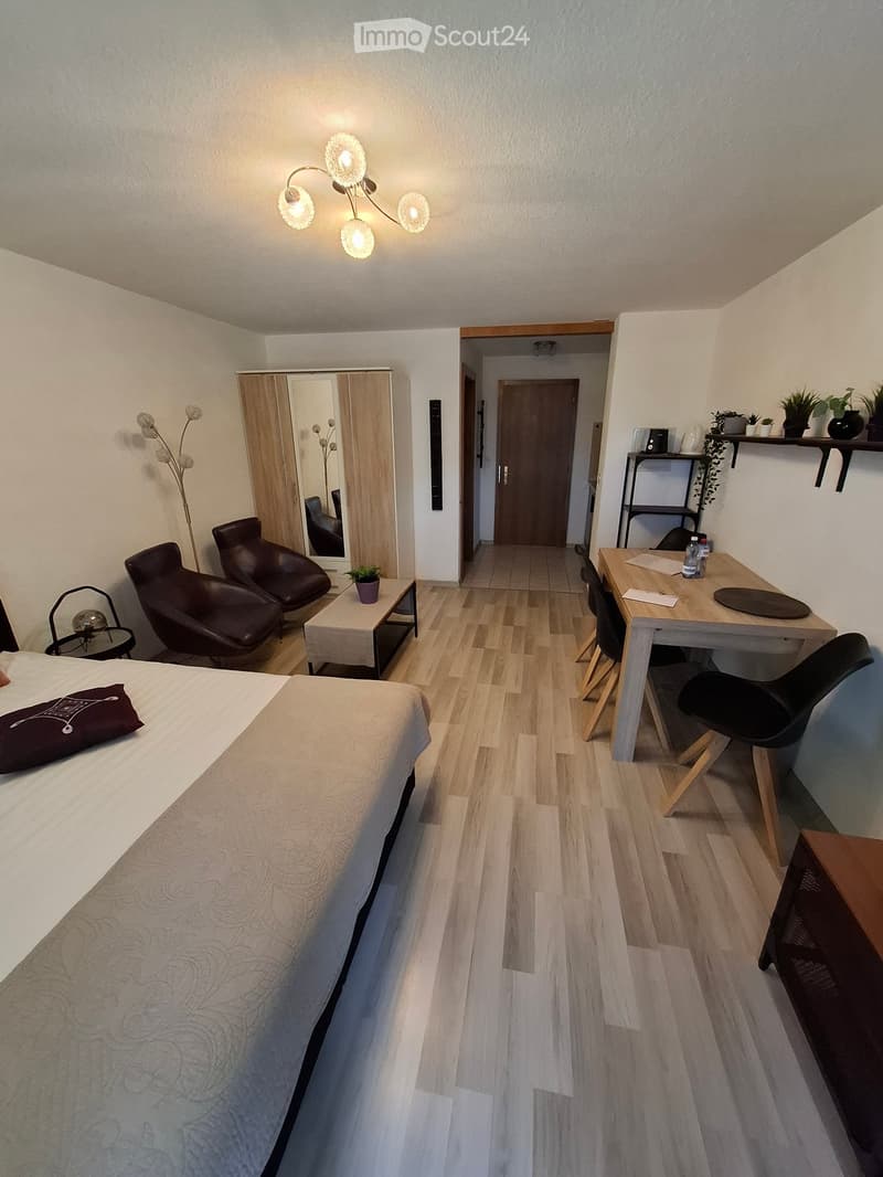 A modern, well-lit bedroom with a large bed, two chairs, a small table, a mirror, a lamp, and a plant. The room features wooden flooring and a door leading to another room.