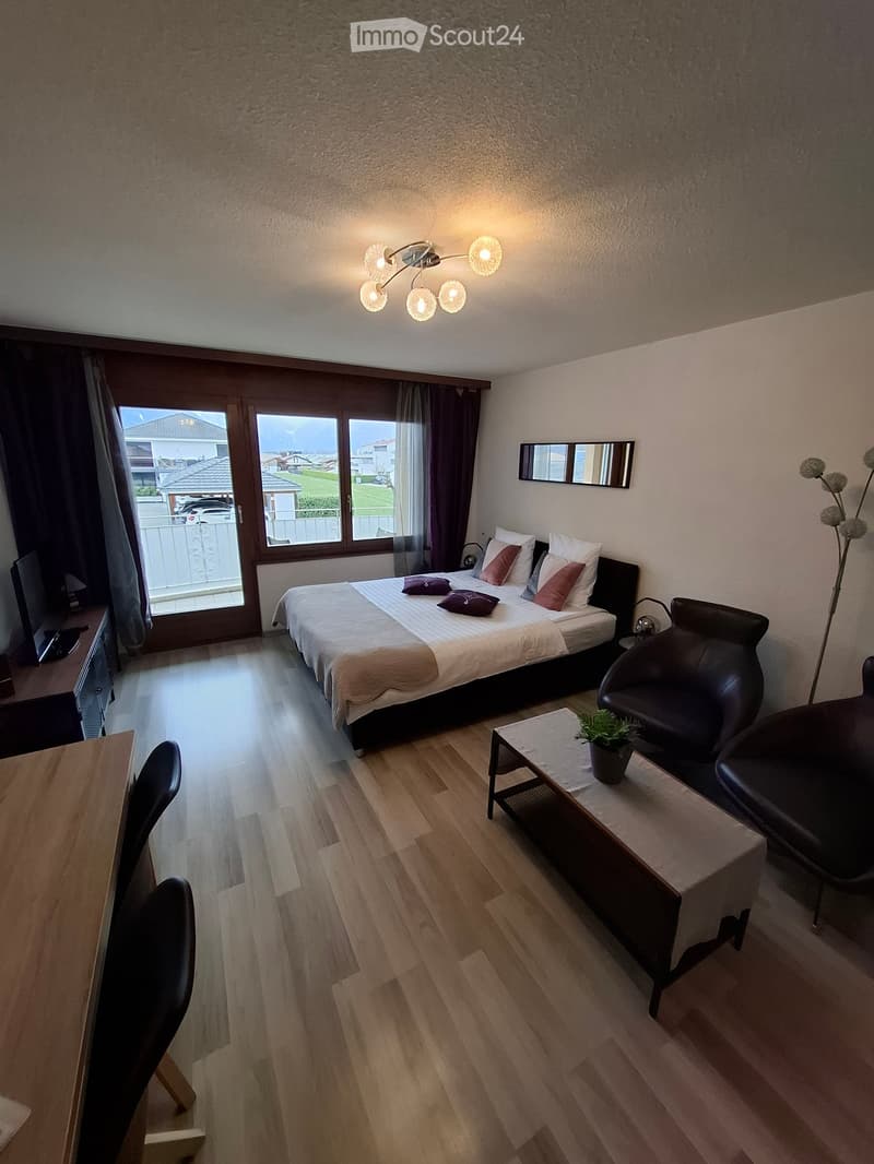 Modern bedroom with a double bed, nightstand, chair, small table, and a large window with a view of the outdoor area.