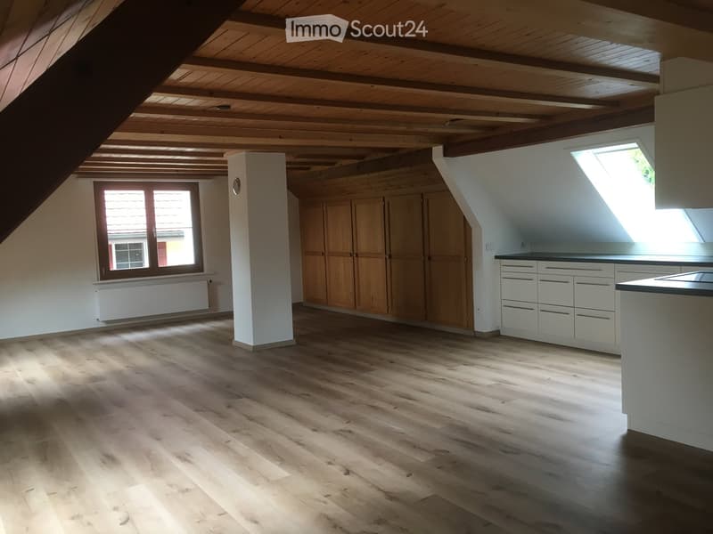 A cozy attic with wooden floor, white ceiling, white cabinets, large window with wooden shutters, built-in wardrobe, ceiling with wooden rafters, skylight