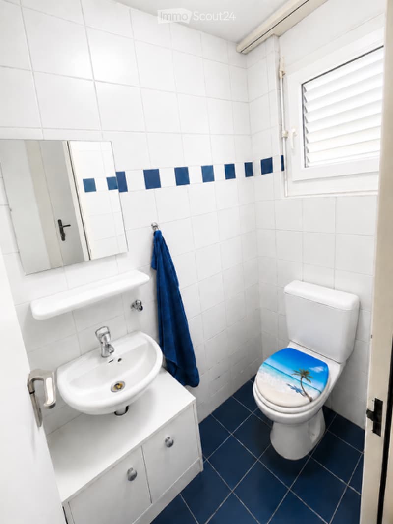 A small, modern bathroom with white tiled walls, a white sink with a mirror, a toilet with a beach-themed seat cover, and a blue towel hanging on the wall.