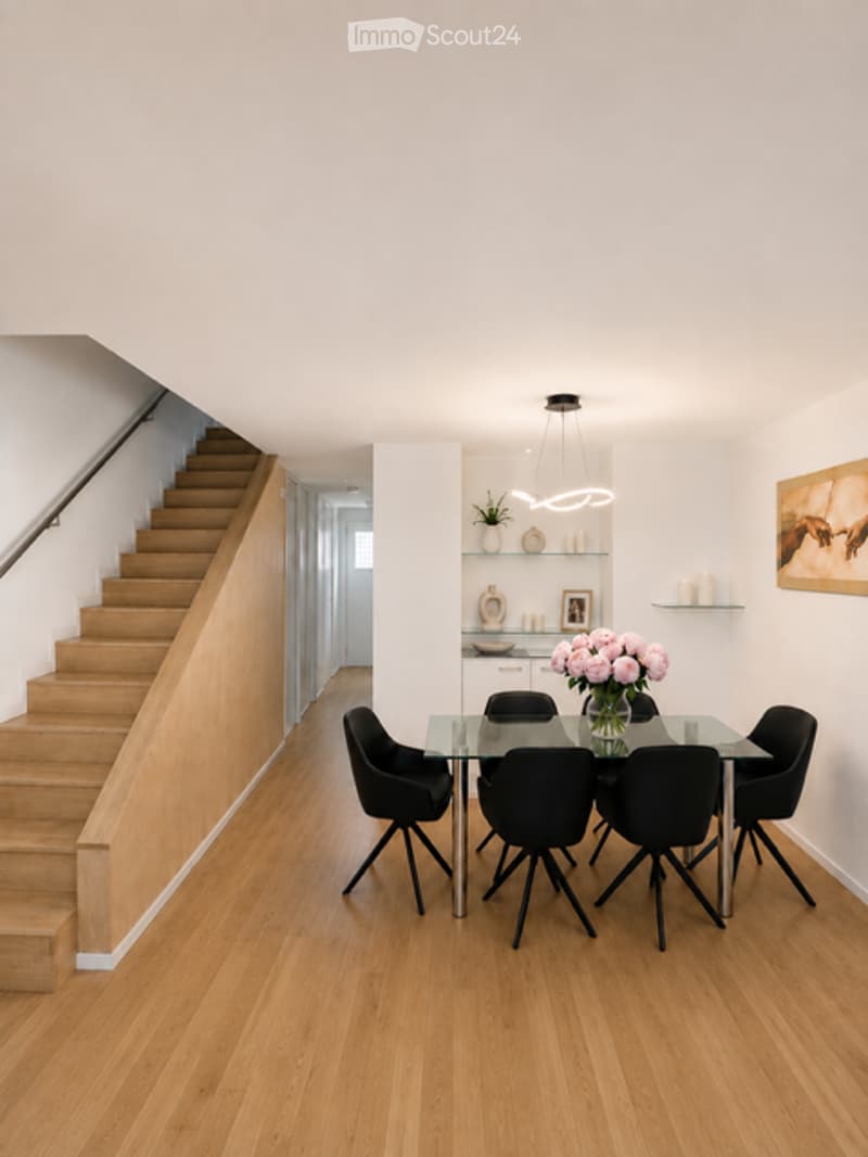 Modern dining area with a glass dining table, black chairs, and a small potted plant on a shelf. The room features light wooden flooring and a staircase leading upwards.