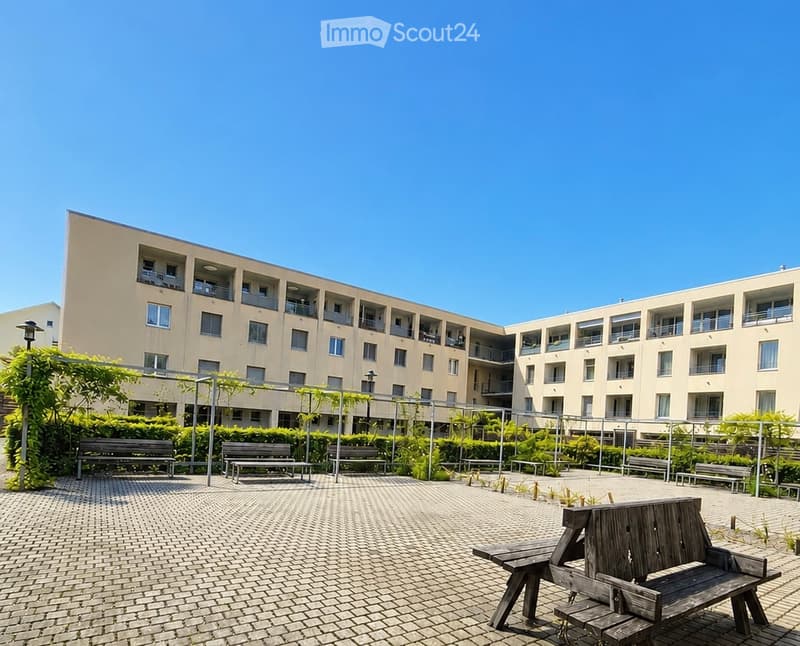 Courtyard area with multiple benches, surrounded by a modern apartment building with balconies and windows.