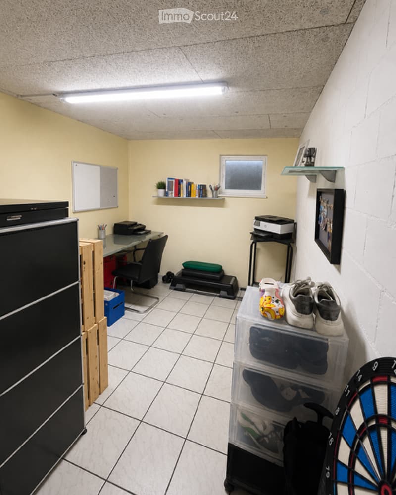 A small, furnished room with a desk, chair, black fridge, shelves with books and a plant, storage bins, and a dartboard on the floor.