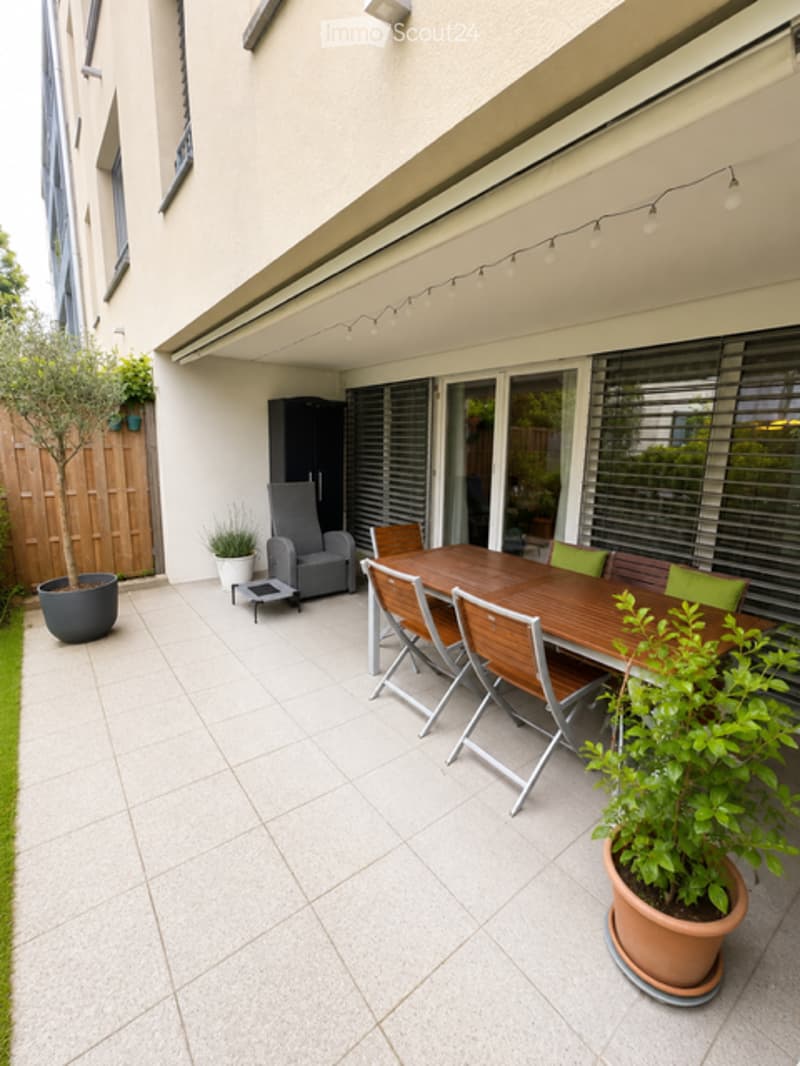 A modern patio area with a wooden dining table, chairs, a grey armchair, and potted plants. The space features large sliding glass doors, string lights, and a tiled floor.