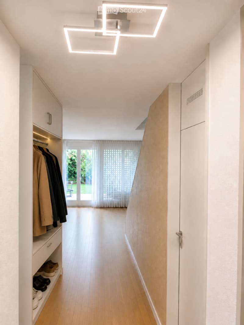 Modern hallway with wooden flooring, built-in storage, and recessed lighting. Features a large window at the end with sheer curtains.