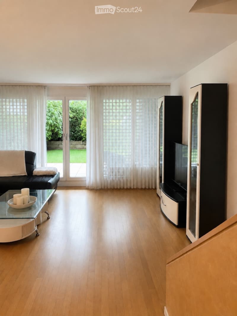 Modern living room with hardwood flooring, large windows with white curtains, a black leather sofa, a glass coffee table, and a TV stand.