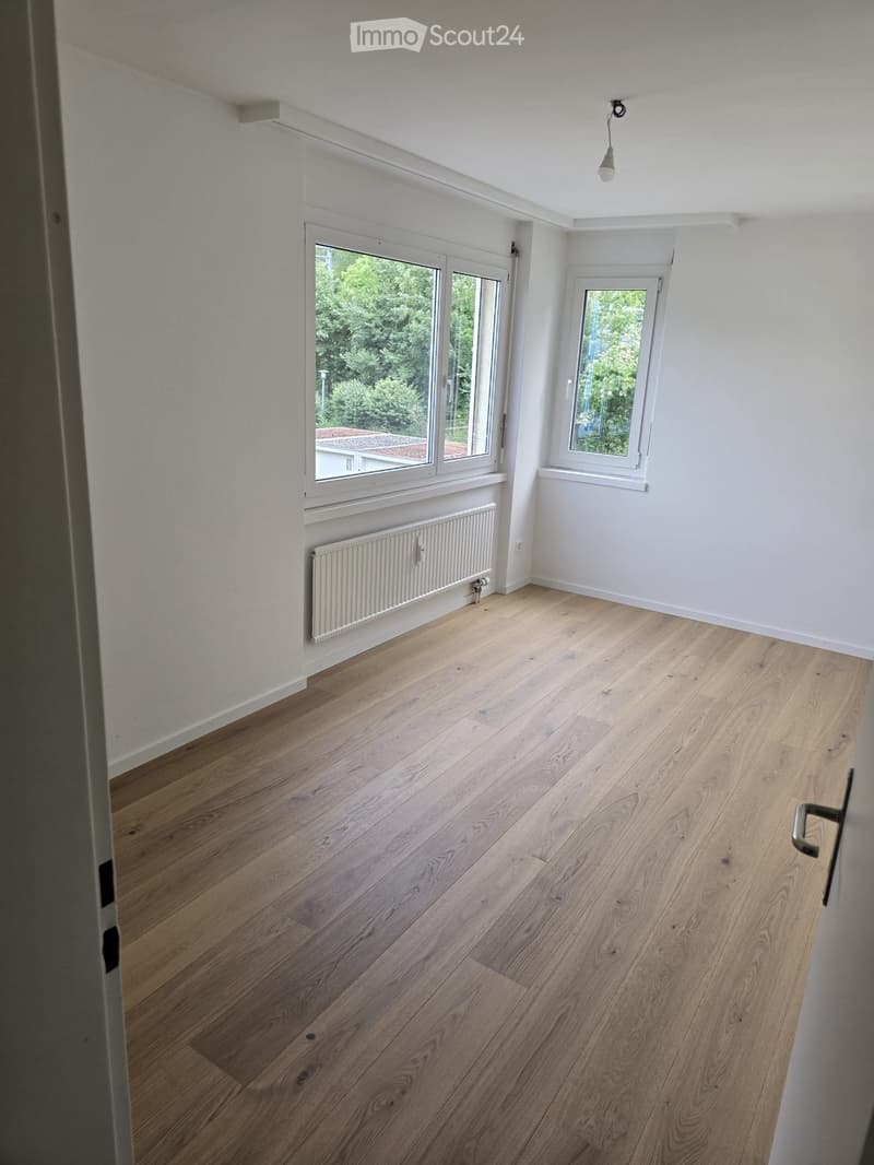 Empty room with light wooden flooring, white walls, and large windows offering a view of greenery outside. A radiator is visible under one window, and a single light fixture hangs from the ceiling.