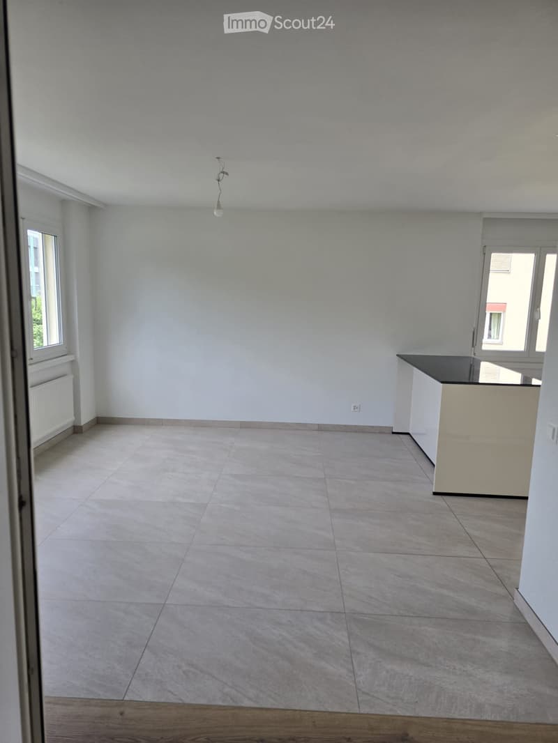 Empty room with light grey walls, tiled floor, and a small kitchen counter in the corner. Windows on two walls allow natural light to enter.