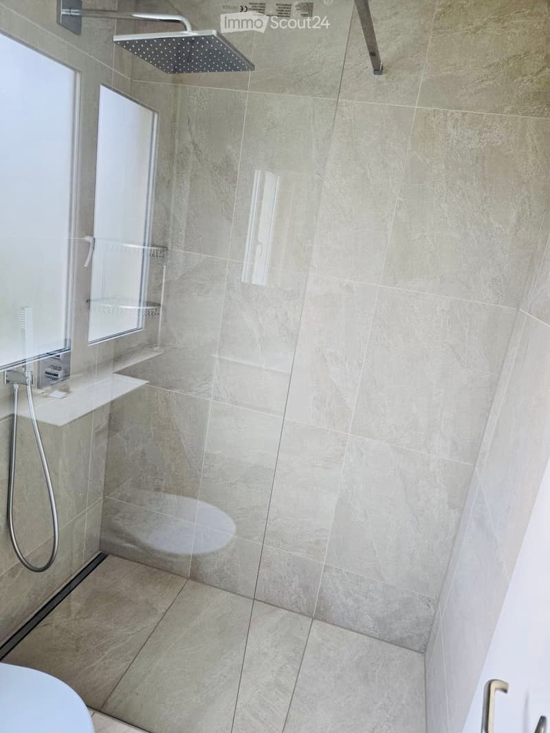Modern bathroom with a glass-enclosed shower area, a large showerhead, and a sleek faucet. The walls and floor are tiled in a light, neutral color.