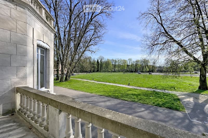 Balcony overlooking a large, open green lawn with trees and a pathway leading to the street.