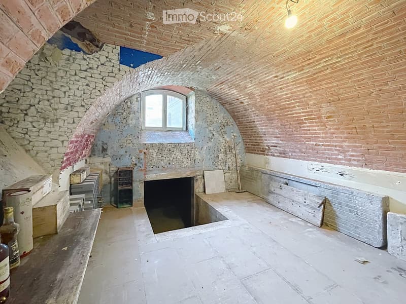 Empty room with stone walls, wooden ceiling, arched doorway, and a small window. Exposed construction materials and tools are visible.