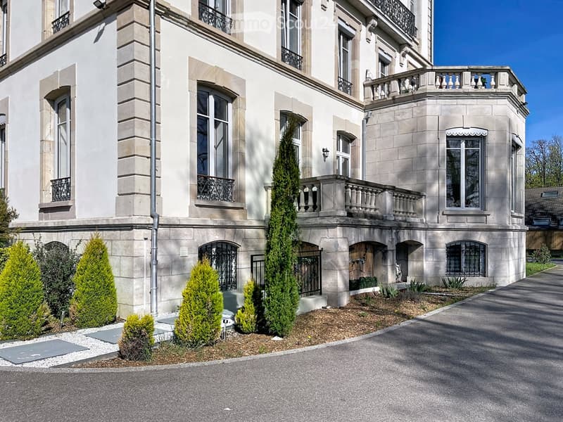 A large, elegant three-story building with beige stone facade, multiple windows, balconies, and decorative railings. The entrance features a gated pathway with neatly trimmed shrubs and trees.