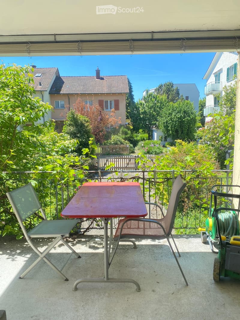 Balcony with a wooden table and two chairs, surrounded by greenery and a metal railing, overlooking a garden and neighboring houses.