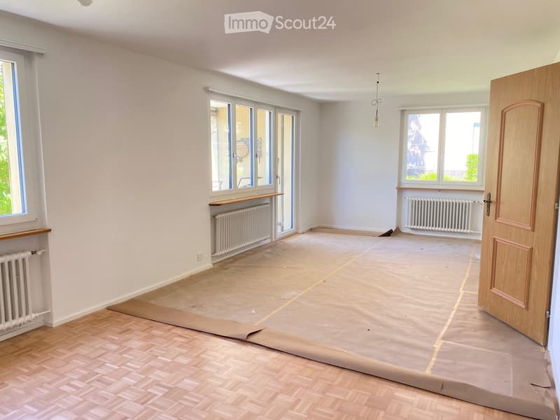 Empty room with light-colored walls, wooden flooring, large windows, radiators, and a partially open door.