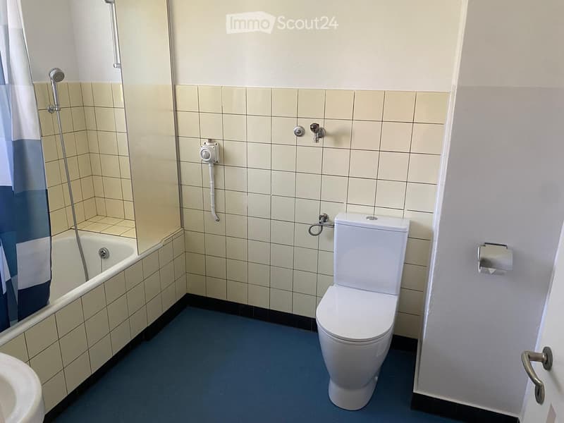 Bathroom with a toilet, bathtub, and tiled walls. Blue floor, white tiles, and a showerhead visible.