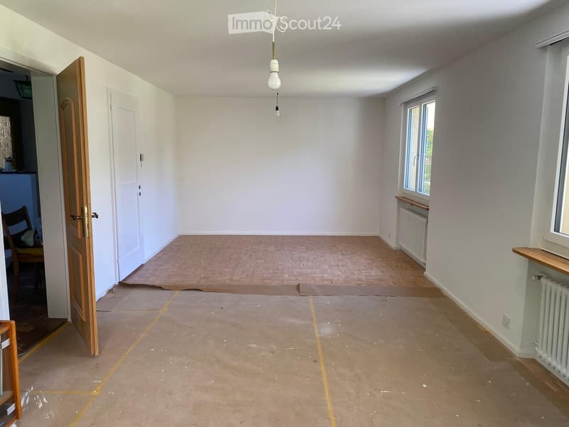 Empty room with light-colored walls, wooden flooring, and a single hanging light fixture. The room has two windows and a door leading to another area.