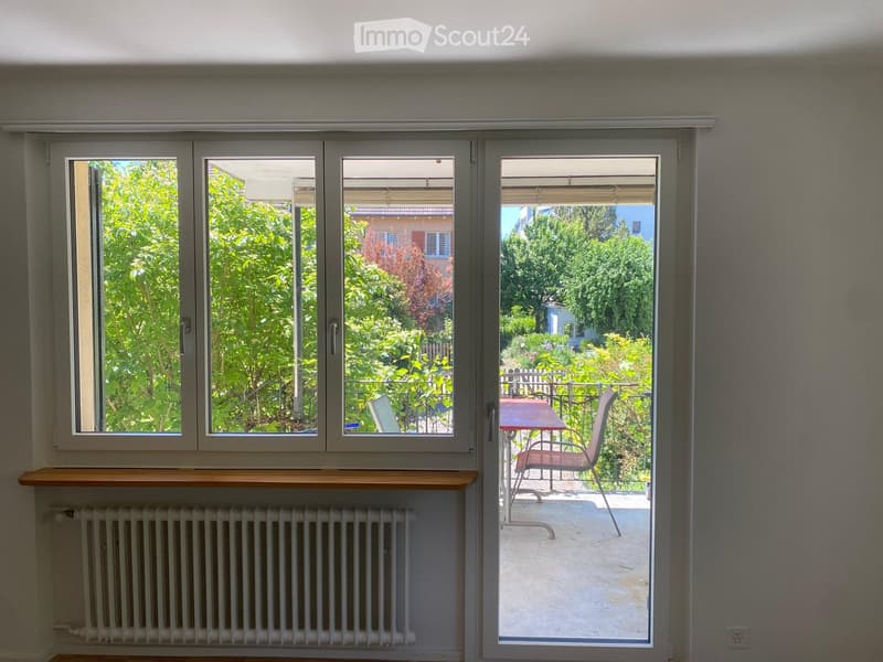 Sliding glass door opens to a balcony with a table and chair, overlooking a green garden area.