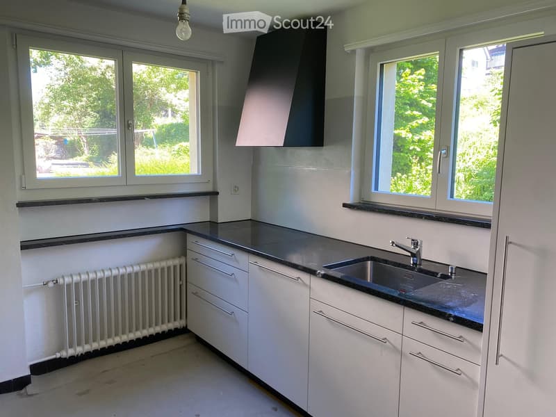 Modern kitchen with white cabinets, black countertops, stainless steel sink, and large windows overlooking greenery.