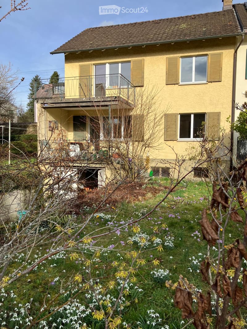 Two-story yellow house with a tiled roof, multiple windows with shutters, and a balcony on the upper level. The front yard features a variety of spring flowers, including snowdrops and daffodils, with some bare branches in the foreground.