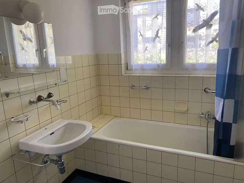 Bathroom with a sink, bathtub, mirror, glass shelf, and handrail. The room features tiled walls and a window with decorative curtains.