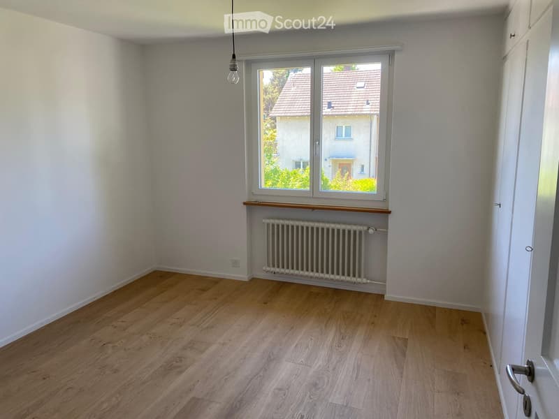Empty room with wooden flooring, white walls, a window with a radiator below, and a hanging light fixture.