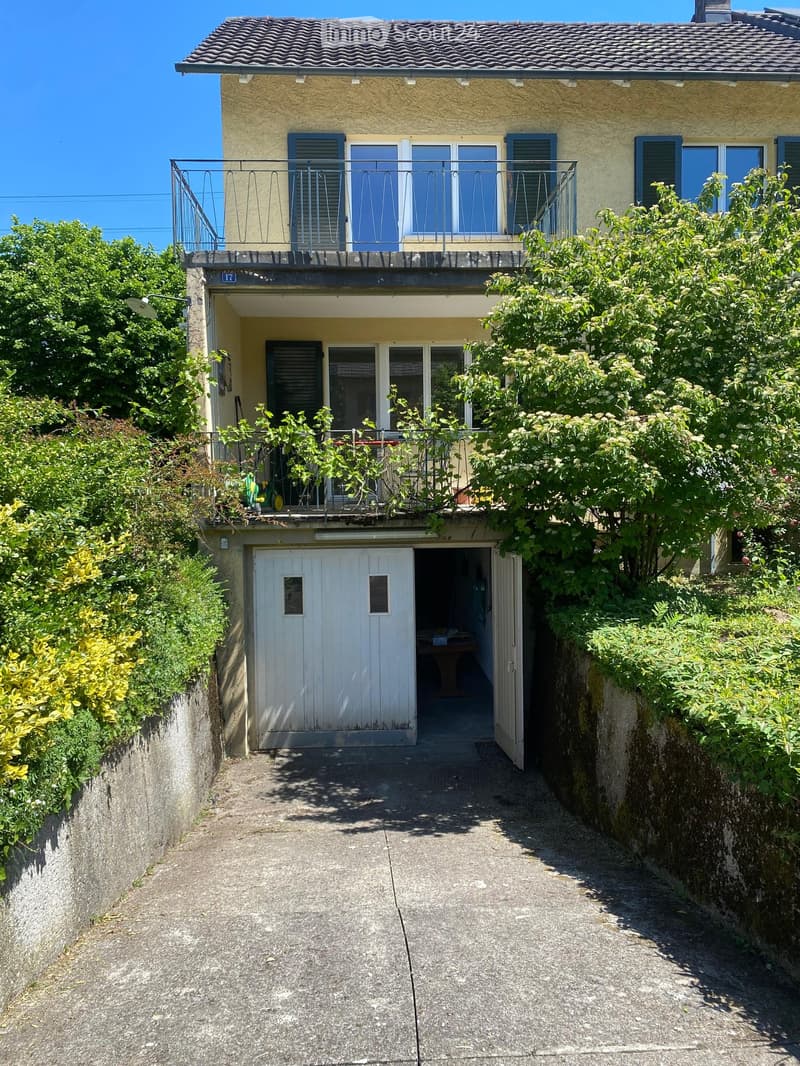 Two-story house with a balcony, garage, and lush greenery surrounding the entrance.
