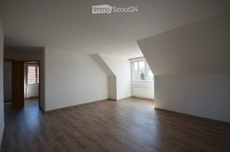 Empty room with wooden flooring, white walls, and large windows allowing natural light. The room features a slanted ceiling typical of an attic space.