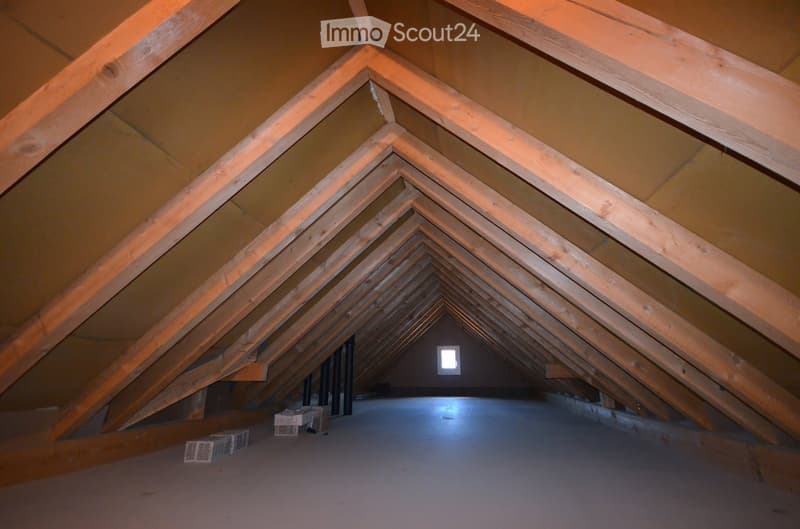 Empty attic space with exposed wooden beams and a small skylight window at the far end. The floor is concrete, and there are a few stacked boxes on the left side.